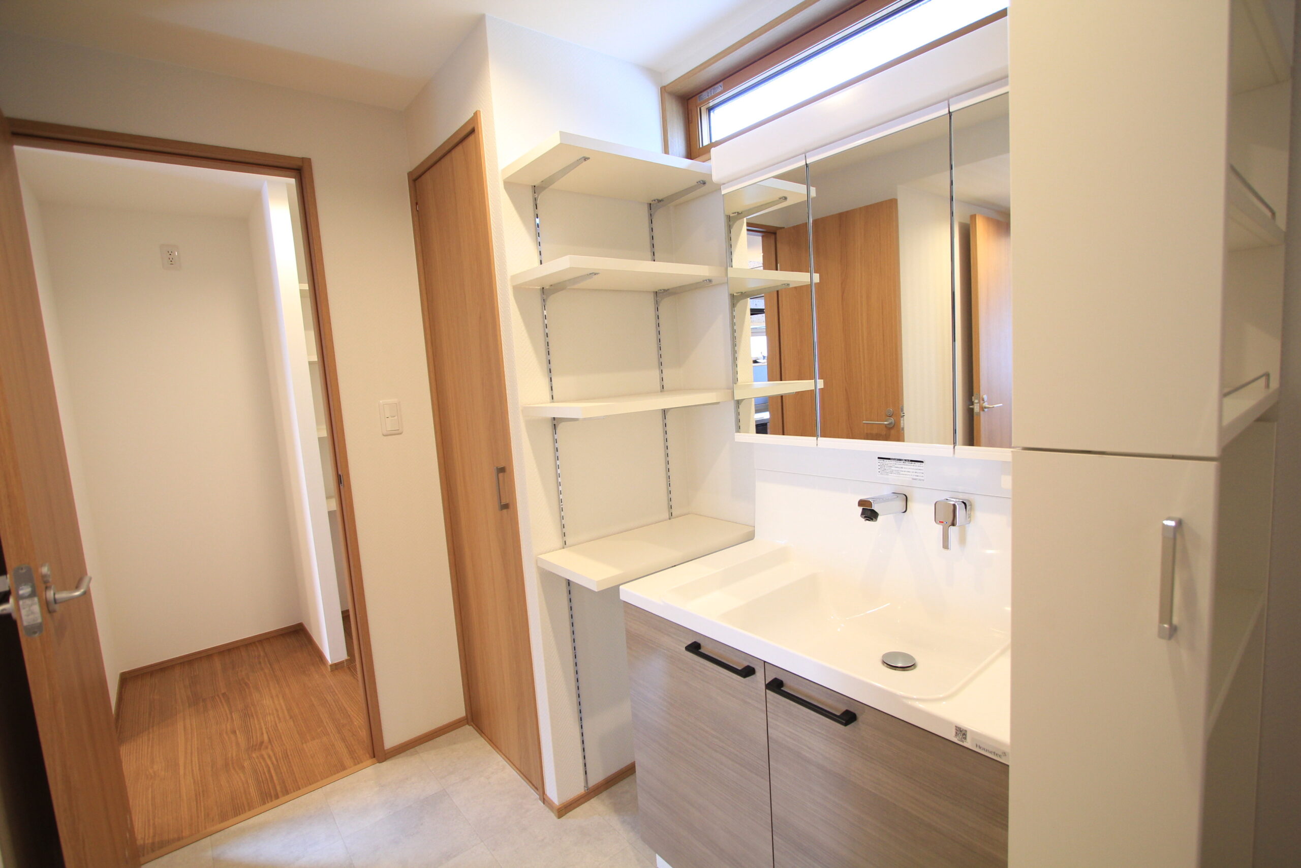 洗面：脱衣室完成写真：収納棚が付いています。（ハイグレードタイプ）奥はキッチンへと繋がります